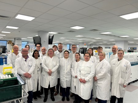 An image displaying a group of people in a laboratory wearing white lab coats. The area they are standing in is surrounded by equipment that is used to test samples such as blood.