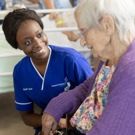 A nurse kneels by the side of an elderly patient, holding their hand, smiling as she tells the nurse a story.