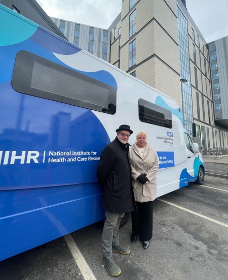 The mobile research hub, which is white with different shades of blue spotted across it, is parked outside the Royal Liverpool University Hospital. Bryn Williams a long-time research participant and Jules West, Associate Director of Research and Innovation at UHLG, stand in front, smiling, during its official unveiling.