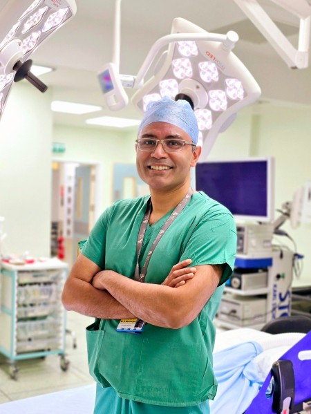 Mr Rajarshi (Rishi) Mukherjee, Consultant Surgeon in Emergency General and Major, wearing his green scrubs and surgical cap, smiling with his arms crossed in theatre in front of a surgical bed.