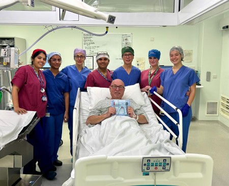 Seven members of the Urology Surgical team line up in their scrubs behind patient, Lee Patten, who is smiling whilst lying in his hospital bed in theatre, holding the Circ Curer staple device.